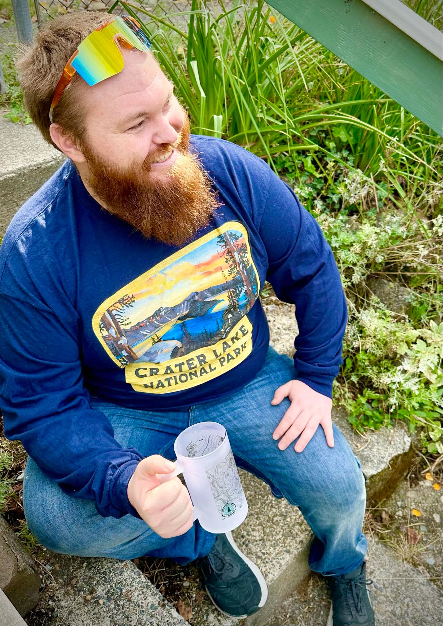 Man wearing a blue sweatshirt with Crater Lake National Park design, holding a mug outdoors.